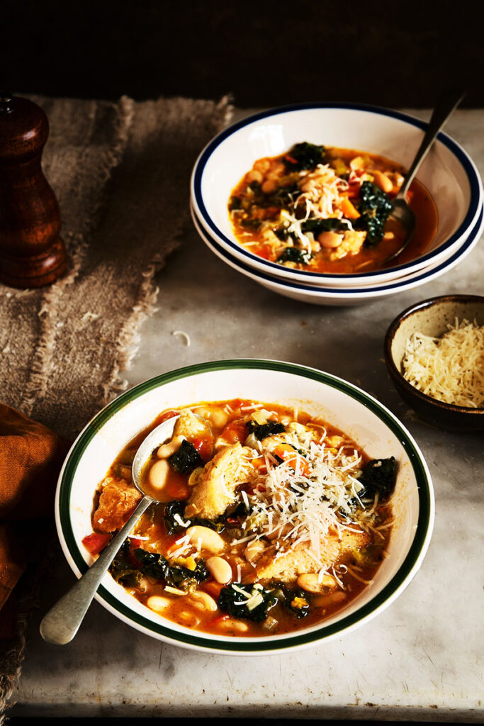 Dark and moody bowls of Ribollita soup with parmesan cheese, peppermill on marble table with textured cloth.